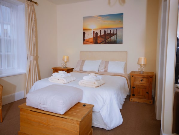 A hotel room with a bed, two nightstands, a wooden bench, a painting of a sunset, and a window with curtains. - The Five Bells (Sandwich)