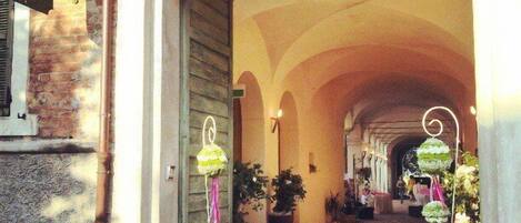 An arched corridor with a brick wall on one side and a wooden door on the other. There are hanging lanterns and potted plants along the corridor.