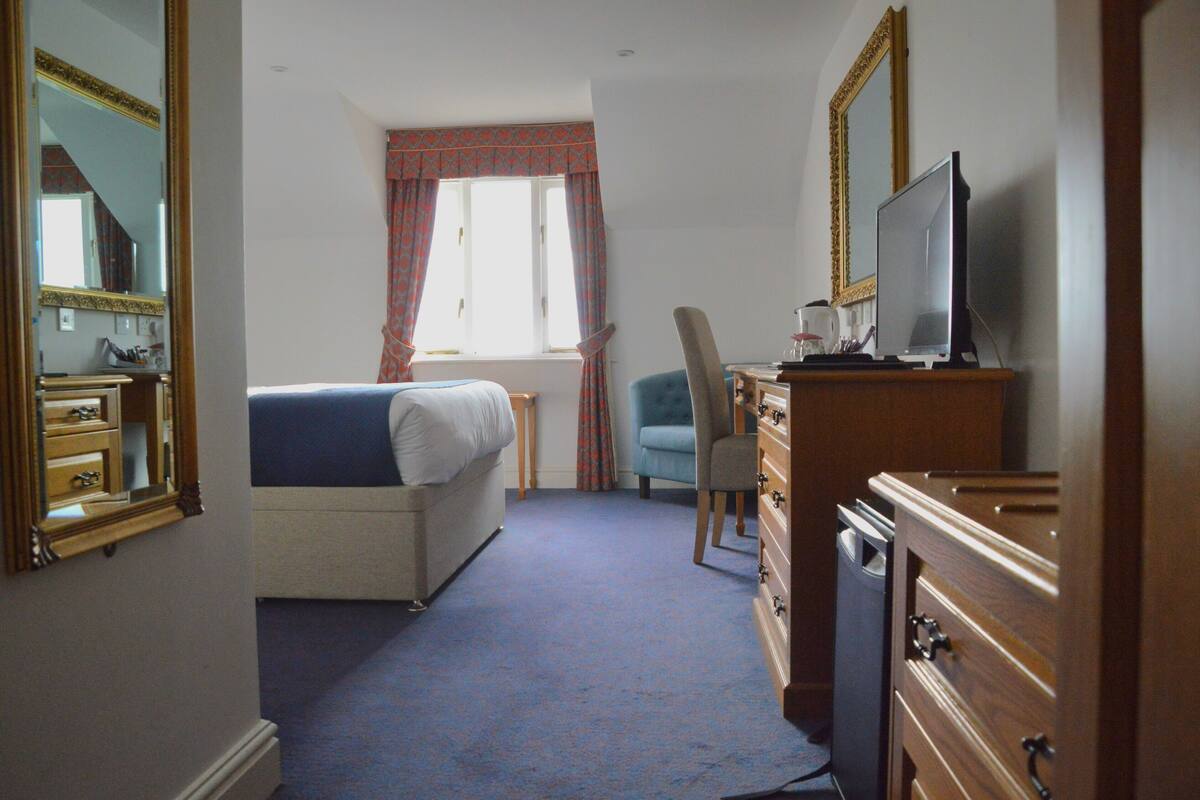 Une chambre d’hôtel avec un lit, une chaise, une commode, un téléviseur et une fenêtre avec des rideaux.