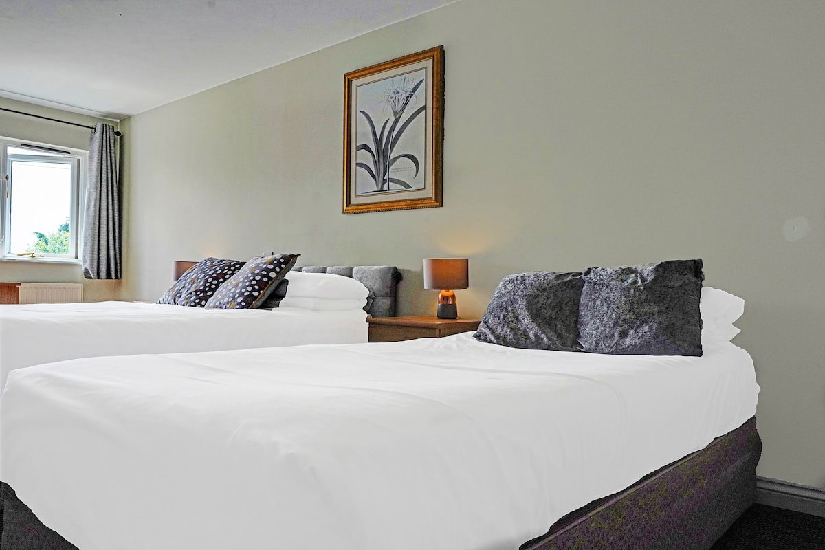 A hotel room with two beds, a nightstand with a lamp, and a framed botanical artwork on the wall.