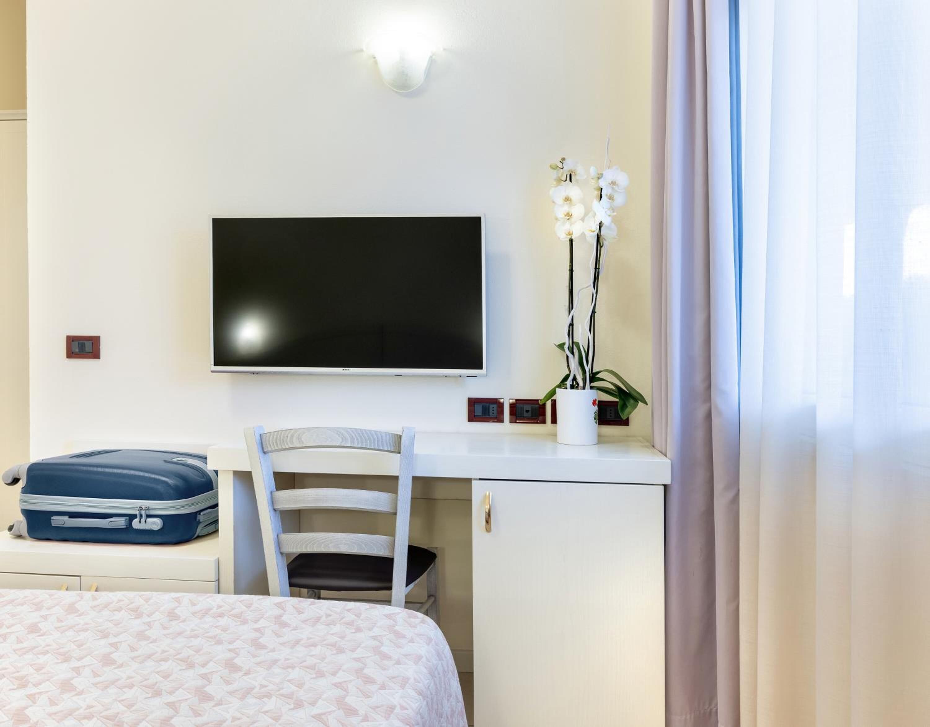 A hotel room with a bed, a desk with a flat-screen TV, a chair, a suitcase, and a potted orchid.