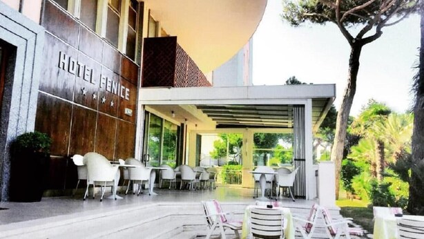 A hotel exterior with a covered outdoor seating area, a sign reading "HOTEL VENICE," and a view of greenery in the background. - Hotel Fenice (Cervia)