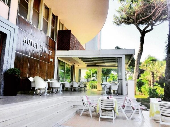 A hotel exterior with a covered outdoor seating area, a sign reading "HOTEL VENICE," and a view of greenery in the background. - Hotel Fenice (Cervia)