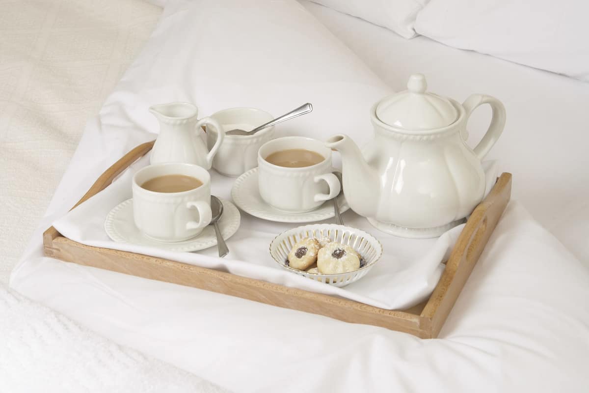 Un lit bien fait, avec du linge de lit blanc et des oreillers, une table de chevet avec une lampe, et un plateau avec un service à thé et des biscuits.