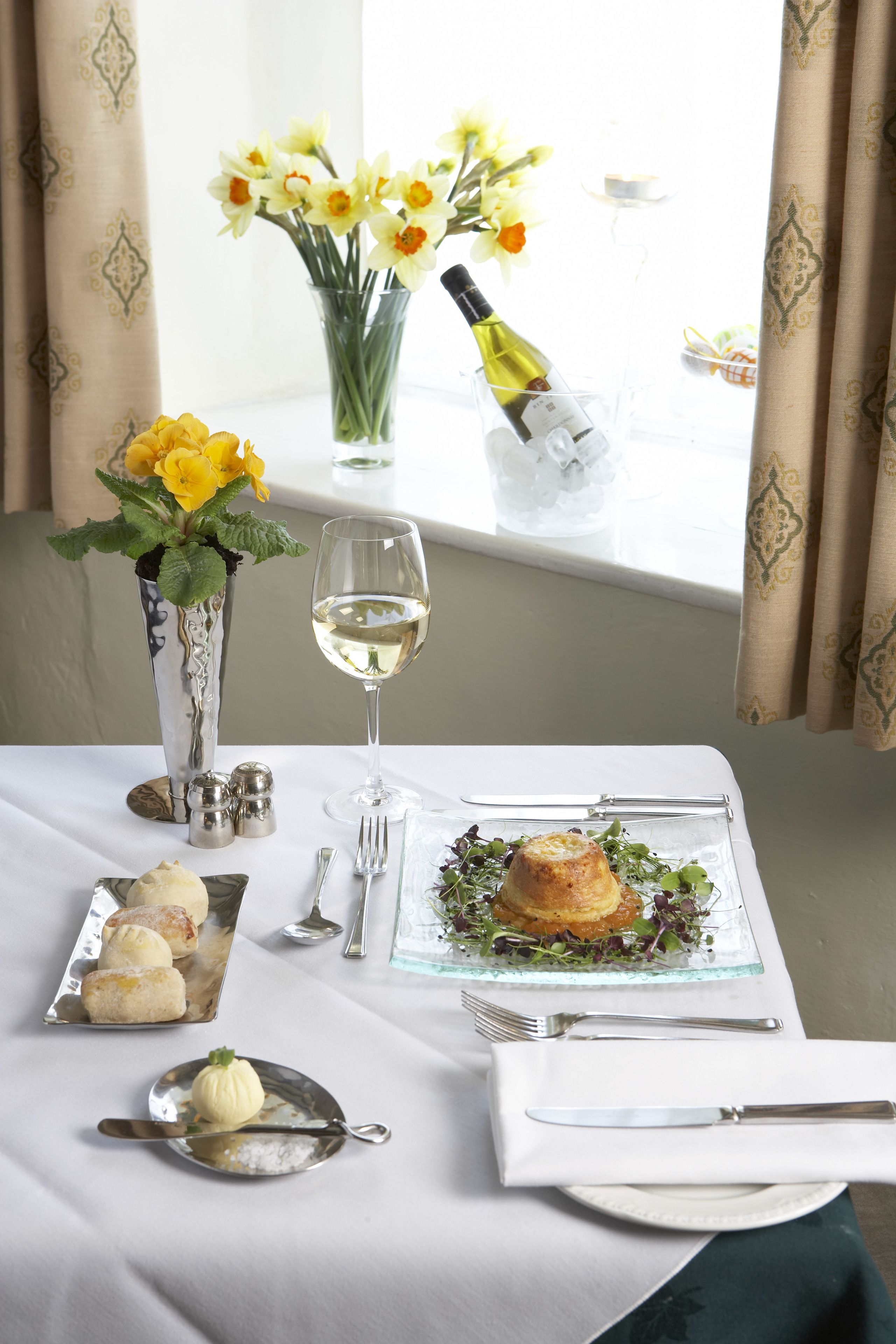 Una mesa de comedor con una copa de cristal para vino, un plato de comida, un pequeño jarrón con flores amarillas y una botella de vino.
