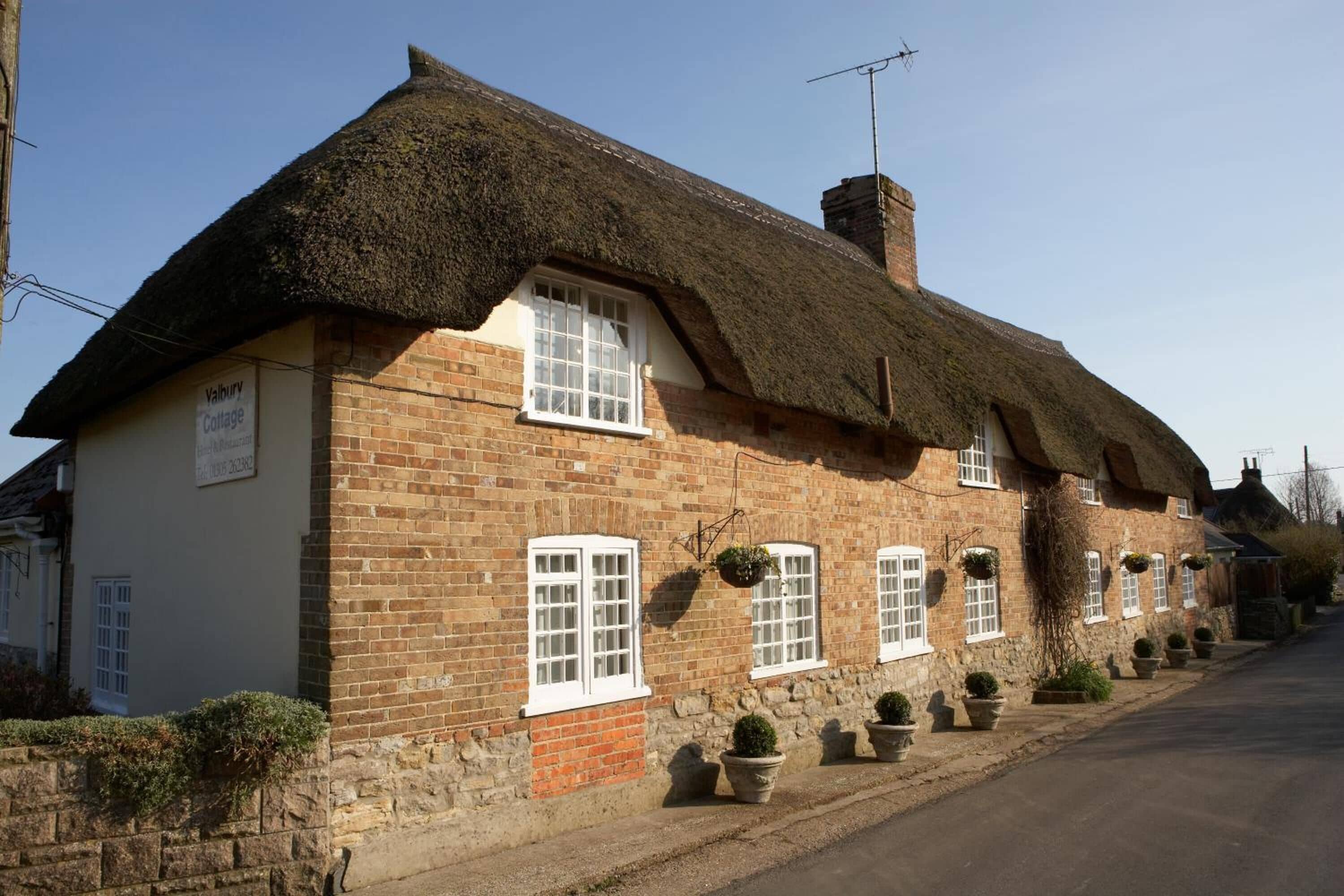 A traditional thatched-roof cottage with a brick exterior, multiple windows, and potted plants along the front.