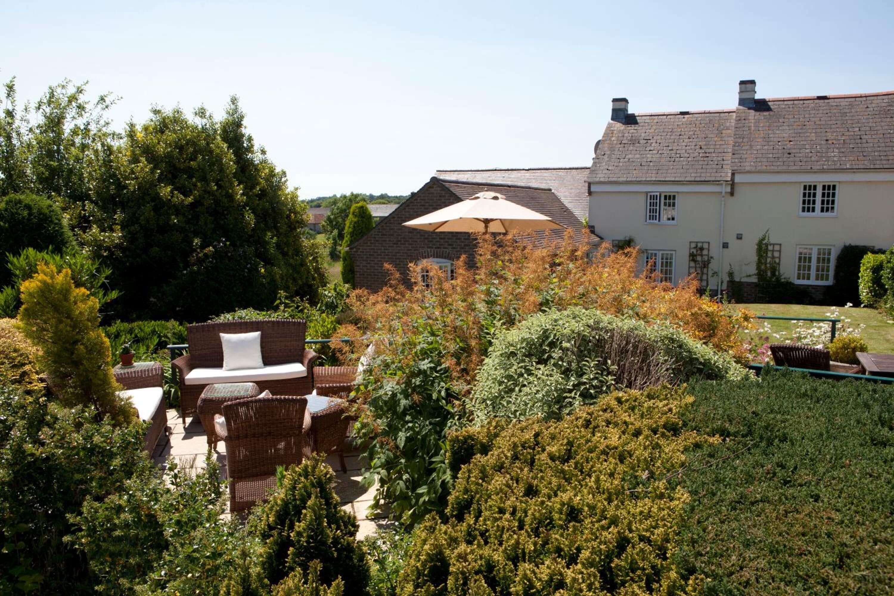 A garden with a wicker sofa and a large umbrella, surrounded by dense foliage and a brick building in the background.