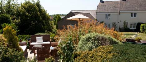 Ein Garten mit einem Korbsessel und einem großen Sonnenschirm, umgeben von dichtem Grün und einem Backsteinhaus im Hintergrund.