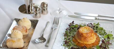 Una mesa de comedor con mantel blanco, una copa de vino blanco, un florero con flores amarillas, un plato de comida y cubiertos.