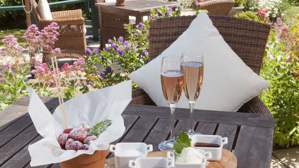Een houten tafel met een fruitmand, twee champagneglazen en kleine witte kopjes op een houten plank.