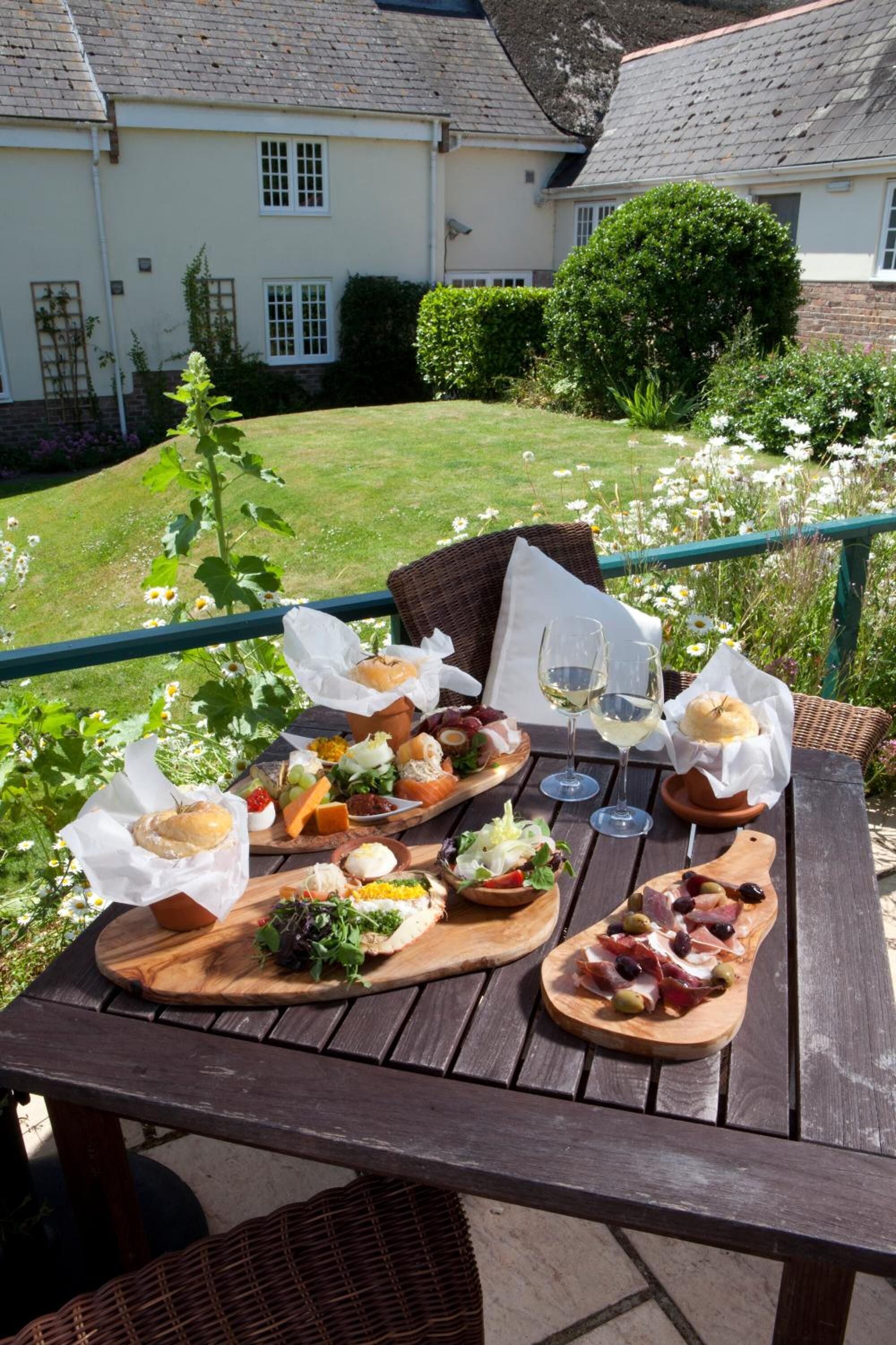 Une table en bois, dressée sur un balcon avec vue sur une maison et un jardin, est garnie de divers plats et de verres à vin.