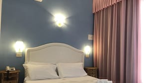 A hotel room with a white bed, two bedside tables, a wall-mounted phone, and a window with pink curtains. - Hotel Negresco (Cervia)