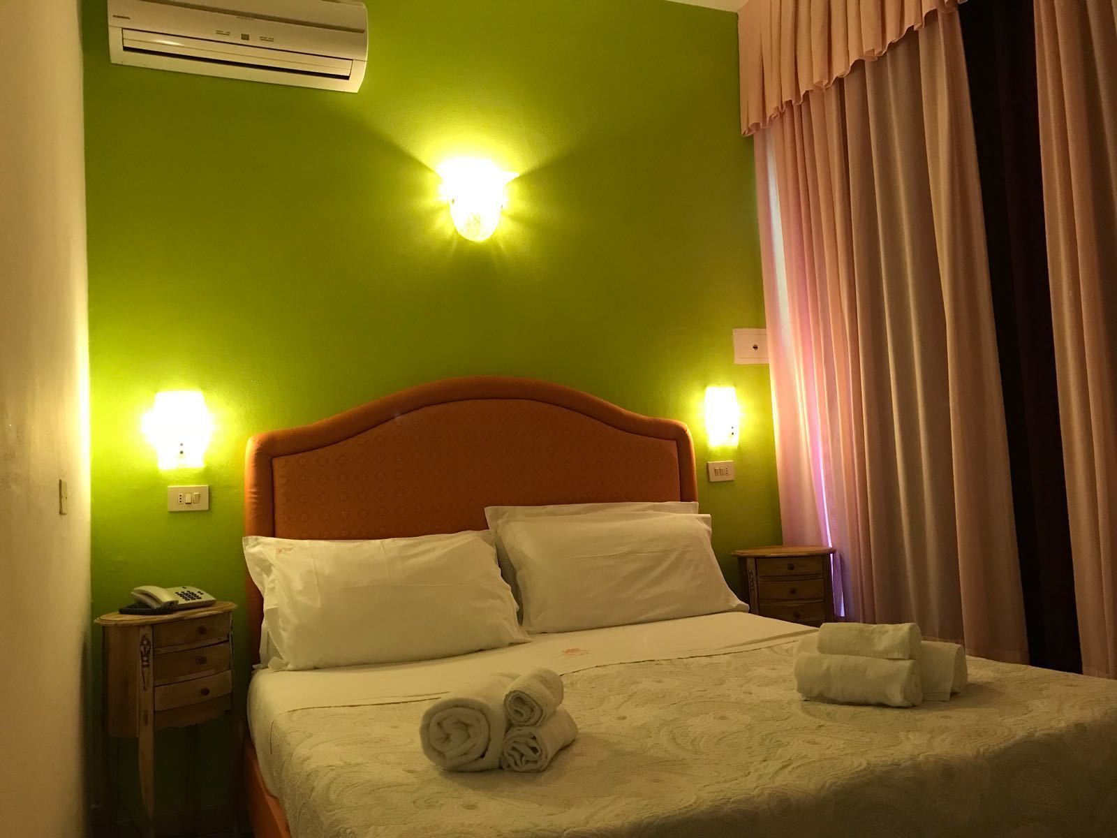 A hotel room with a green wall, a wooden headboard, a bed with white linens, two bedside lamps, a nightstand with a phone, and a window with curtains.