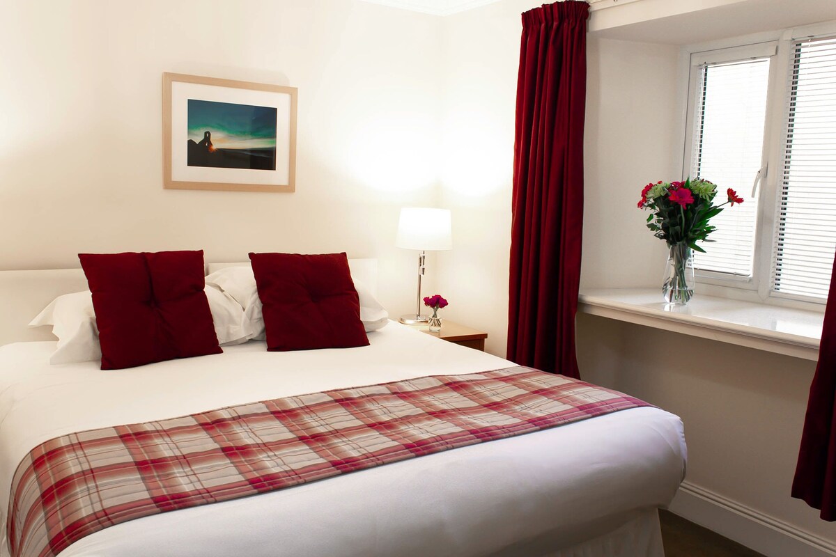 Una cama bien hecha con almohadas rojas, una colcha escocesa, un cuadro en la pared, una lámpara de noche y una ventana con cortinas rojas.