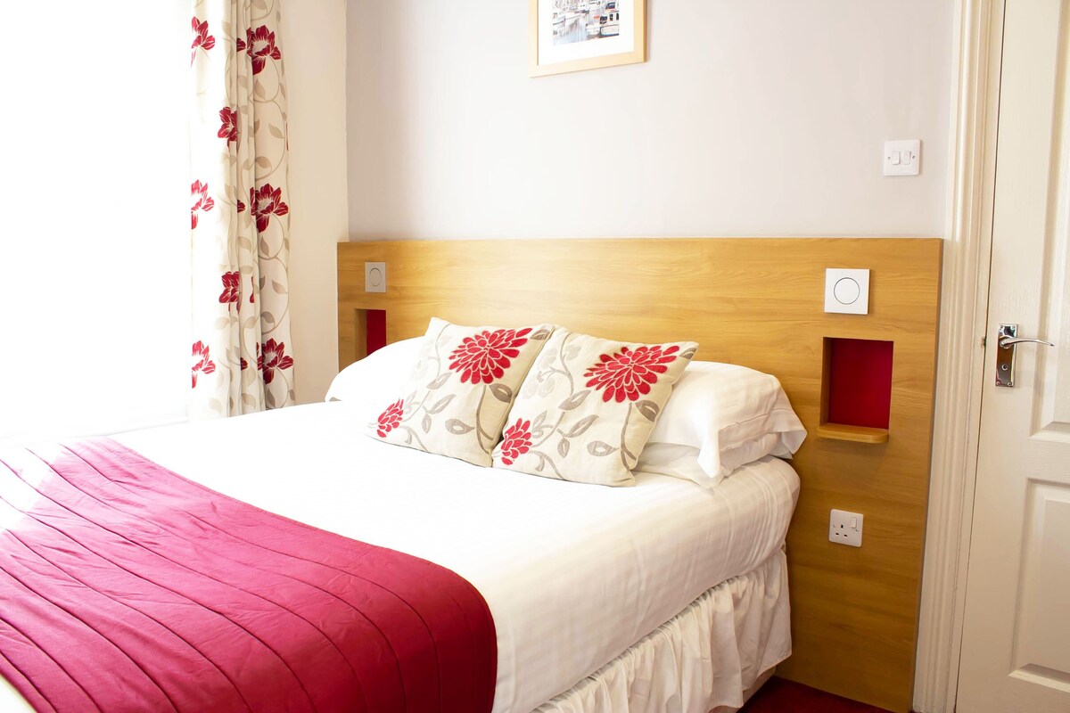 Una cama bien hecha con una colcha roja y blanca, dos almohadas con estampado floral y un cabecero de madera.