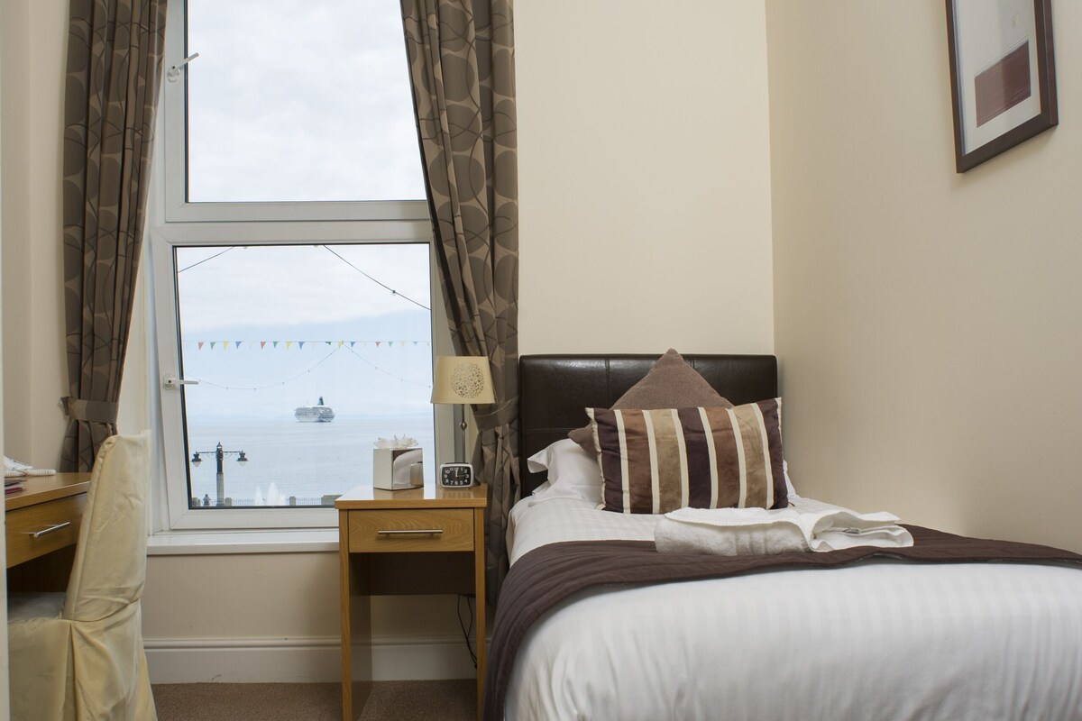 Habitación de hotel con cama, escritorio, silla, ventana con cortinas e imagen colgada en la pared.