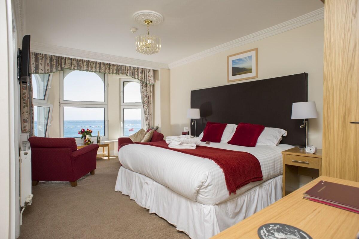 Habitación de hotel con una cama grande, un escritorio, una silla y vista al mar por las ventanas.