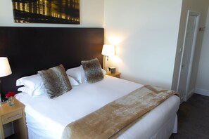 Una cama bien hecha con sábanas blancas, dos almohadas decorativas, un plaid marrón y una mesita de noche con una lámpara.
