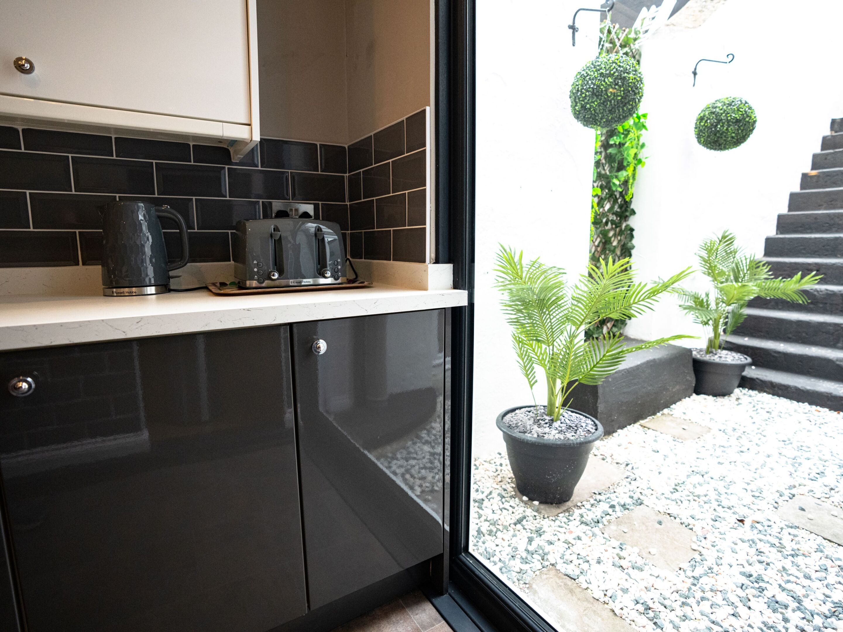 Een moderne keuken met donkere kastjes en een aanrecht, een waterkoker en een broodrooster. Buiten staan potplanten en een trap die naar een balkon leidt.