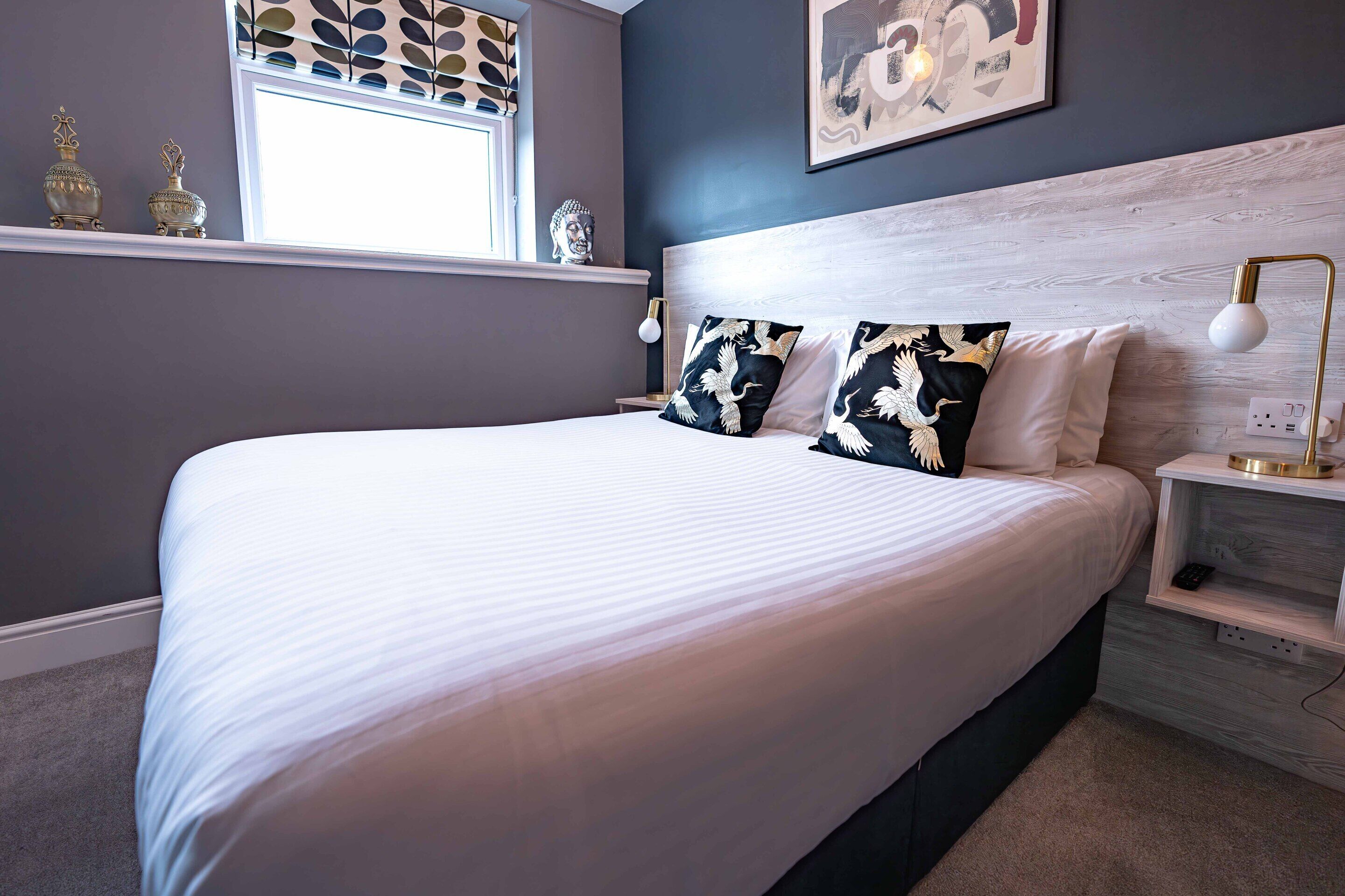 A bedroom with a large bed, a nightstand, a wall-mounted lamp, and decorative items on the windowsill.