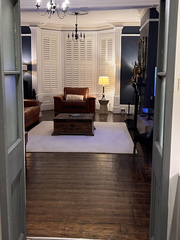 A living room with a brown leather armchair, a wooden coffee table, and a white rug. There is a chandelier above and a lamp on a side table. - Amadeus Apartments (Douglas)