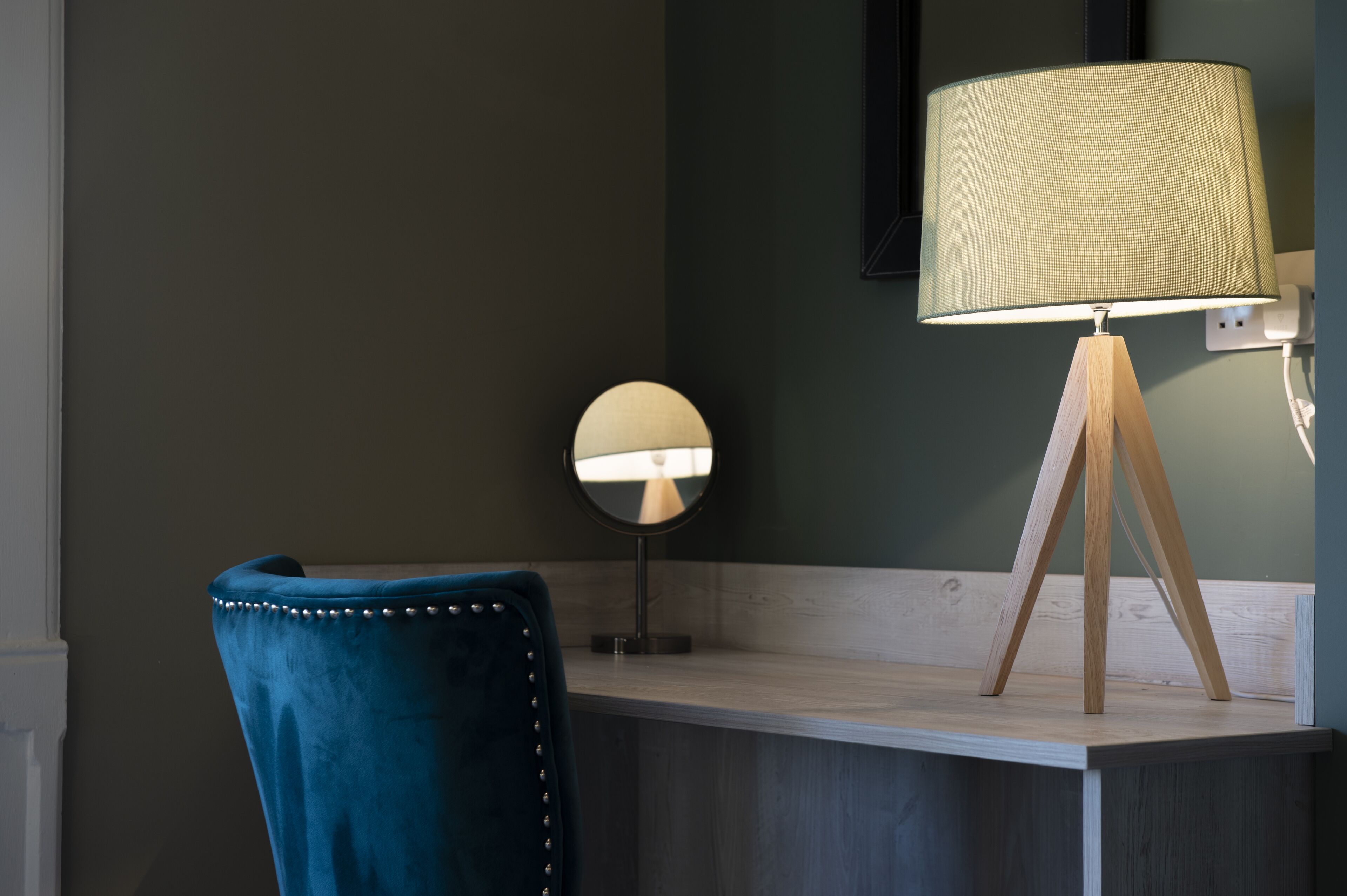 A room with a desk, a round mirror, a blue chair, and a lamp with a wooden stand.