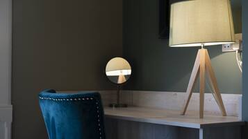 A room with a desk, a round mirror, a blue chair, and a lamp with a wooden stand.