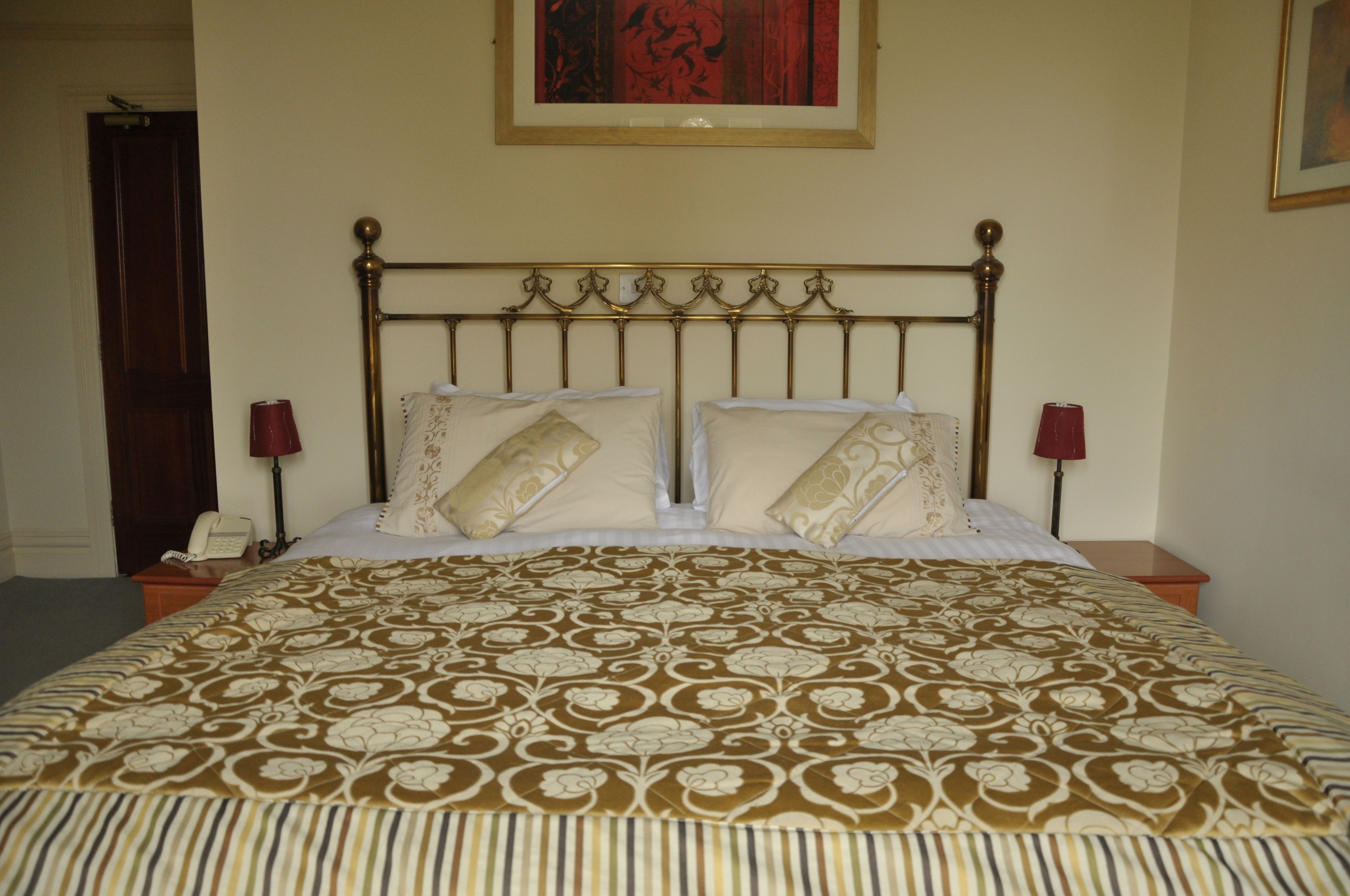 Een net opgemaakt bed met een bedsprei met patroon, twee nachtlampjes en een kunstwerk boven het bed.