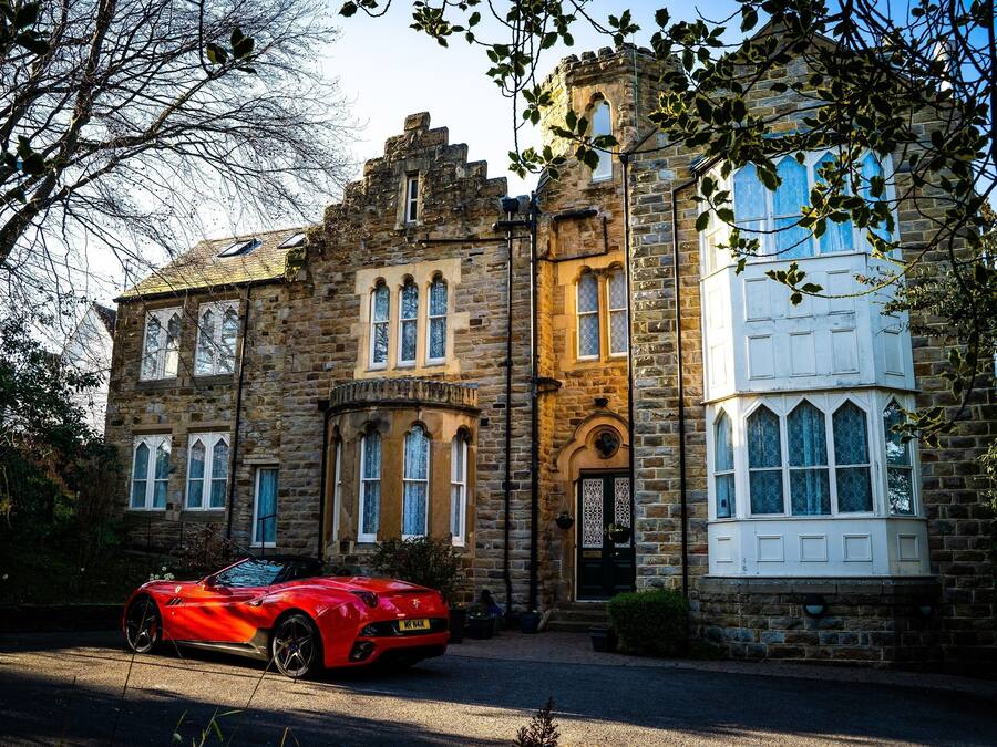 Un edificio histórico de piedra con ventanas arqueadas y una torre, con un coche deportivo rojo estacionado al frente.