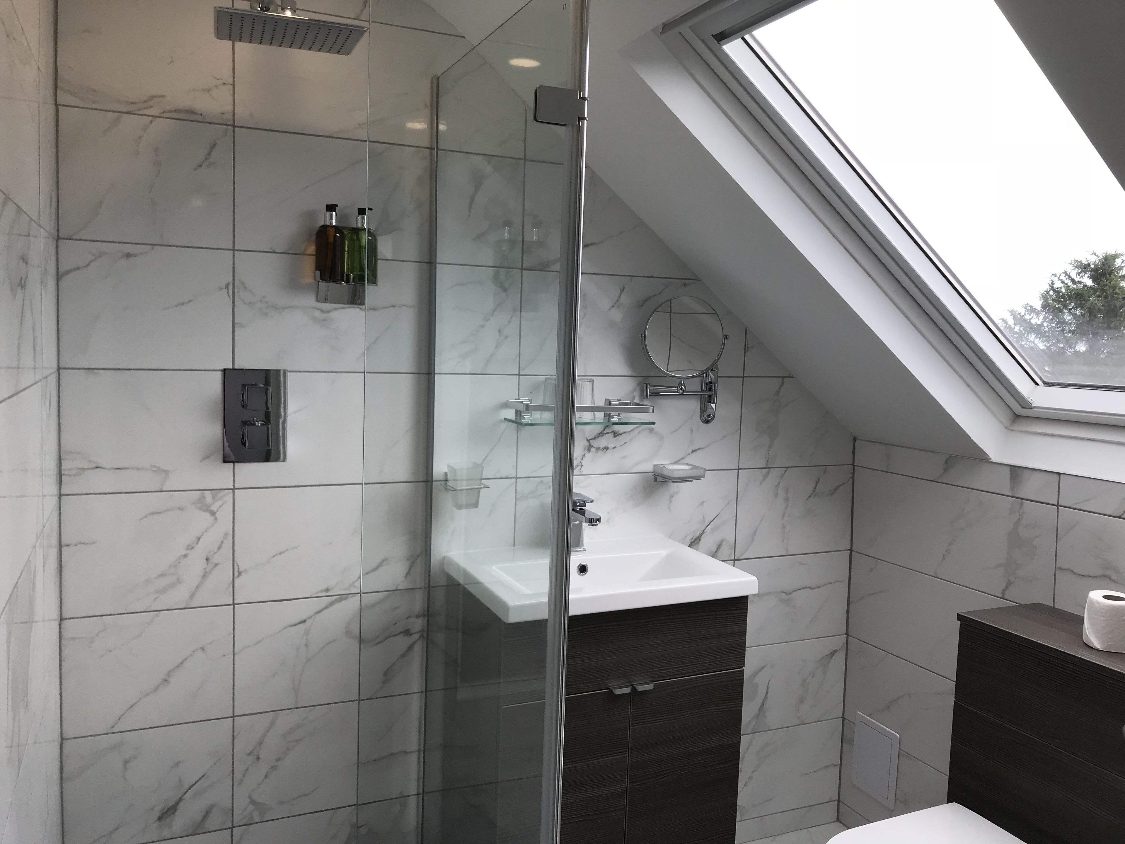 Un baño moderno con ducha envuelta en vidrio, un lavabo blanco y un tragaluz.