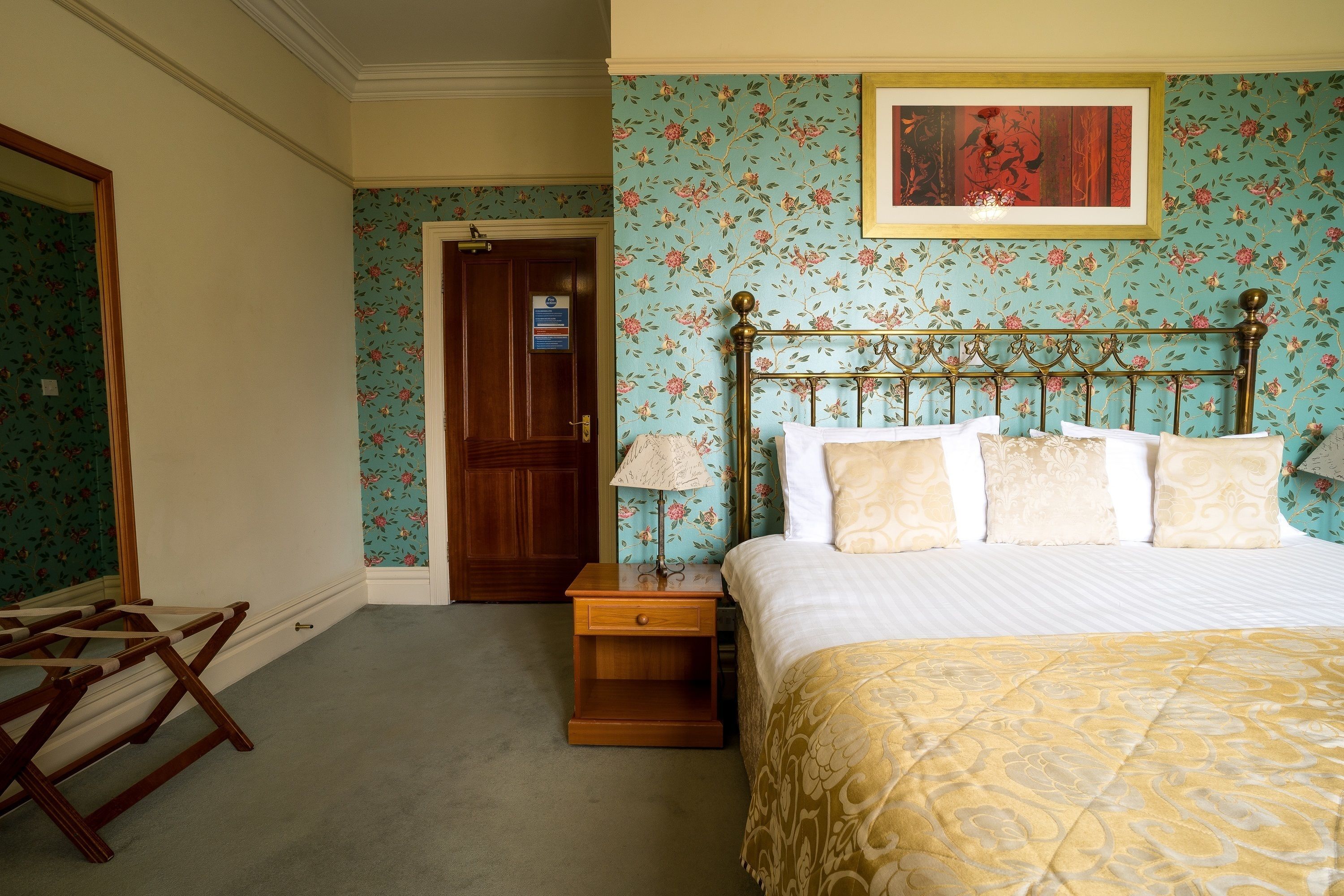 A bedroom with floral wallpaper, a wooden bed frame, a nightstand with a lamp, and a door leading to another room.
