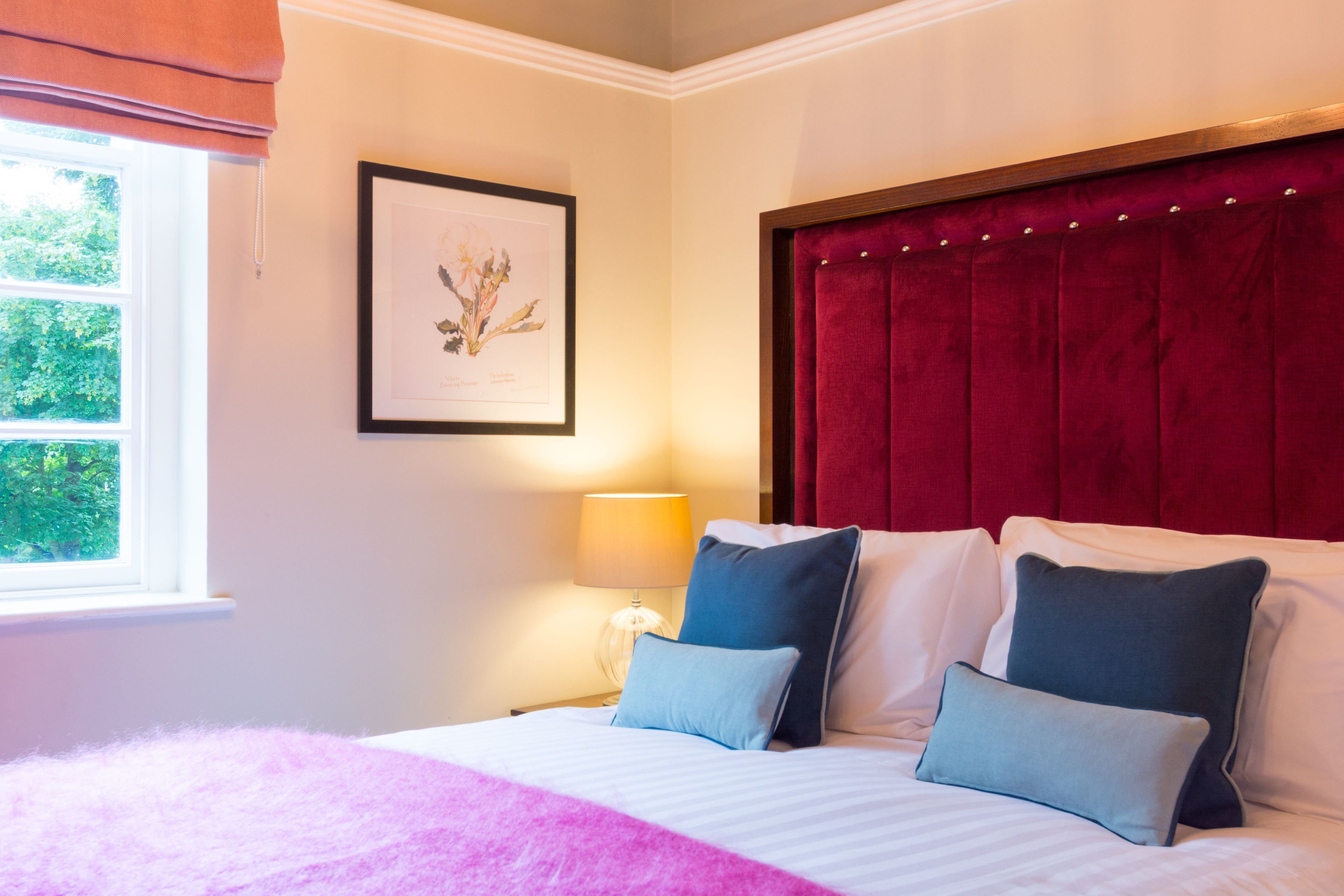 A bedroom with a bed, pillows, a framed picture, a window, and a lamp.