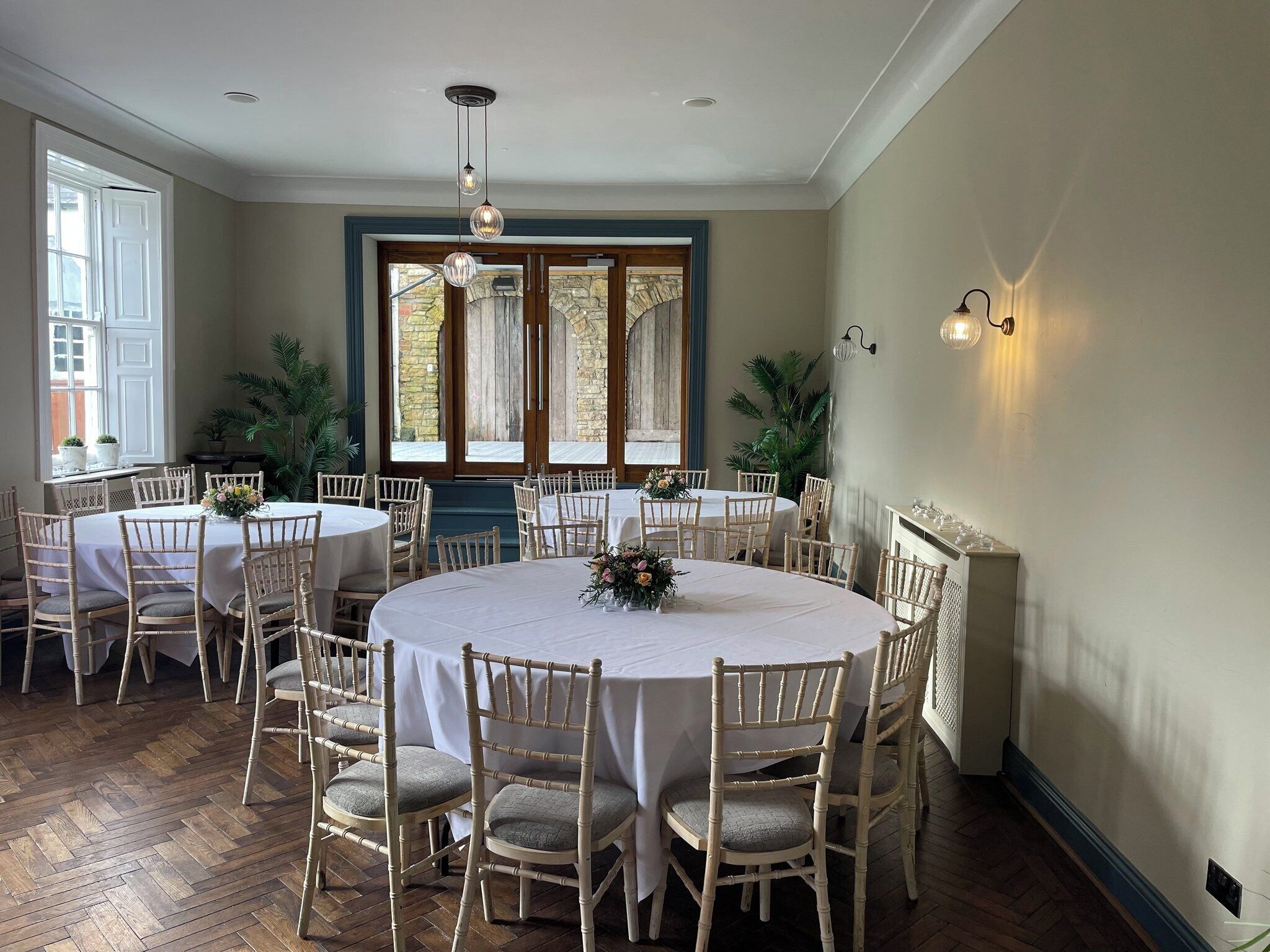 A dining area with round tables and chairs, large windows with curtains, and a stone wall visible through the windows.