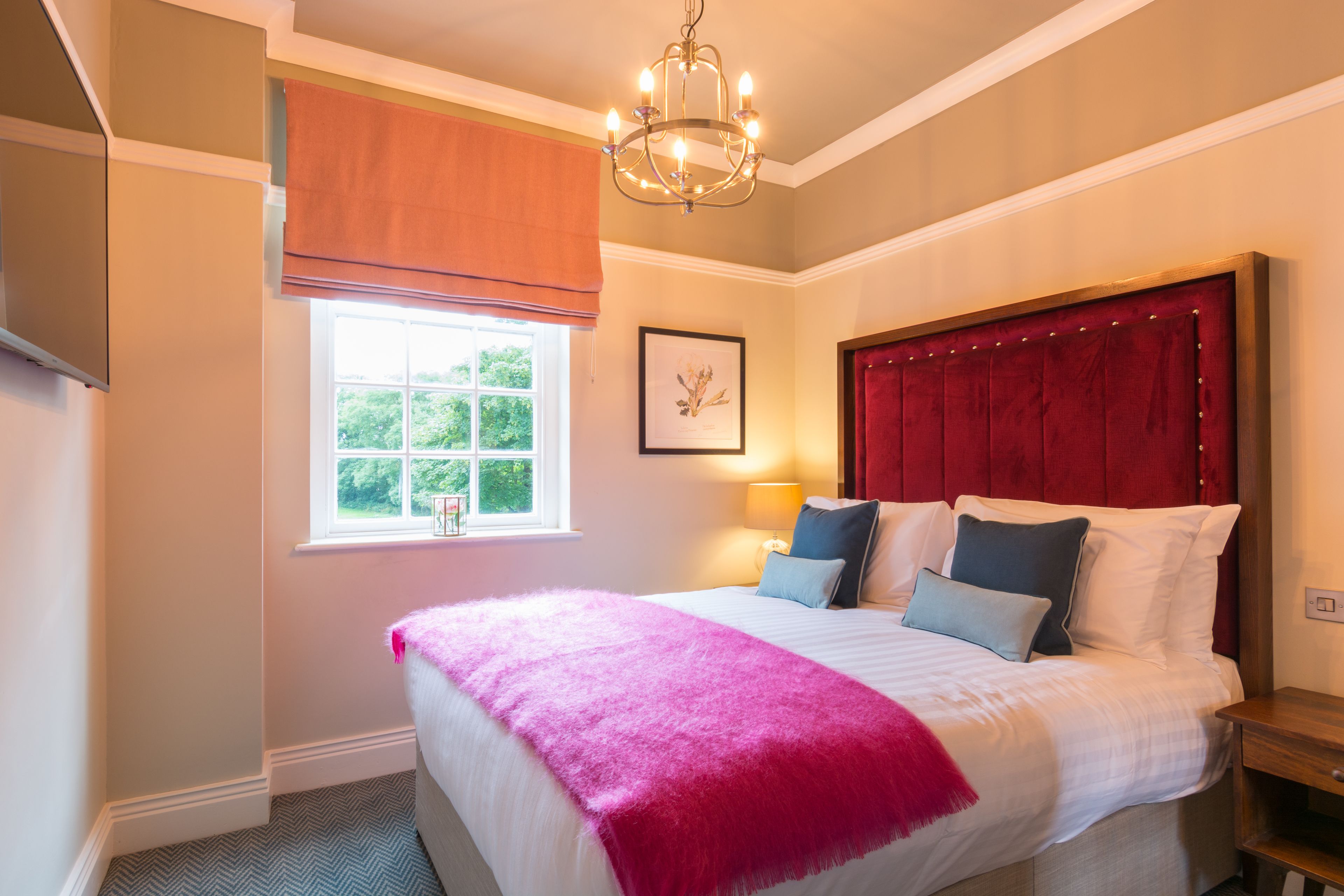A bedroom with a large bed, a red headboard, a window with a view of greenery, and a chandelier.