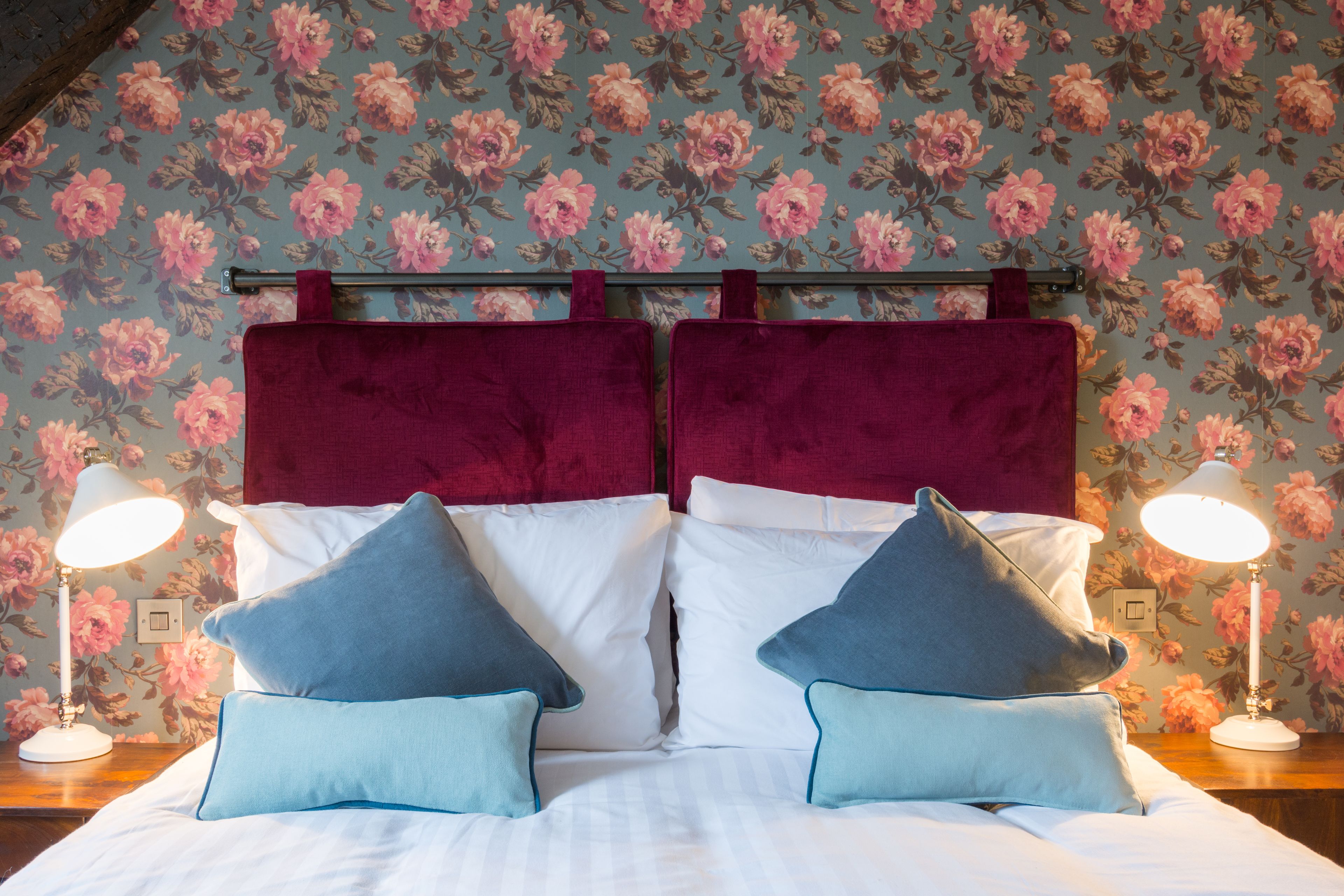 A neatly made bed with white and blue pillows, a red headboard, and floral wallpaper.
