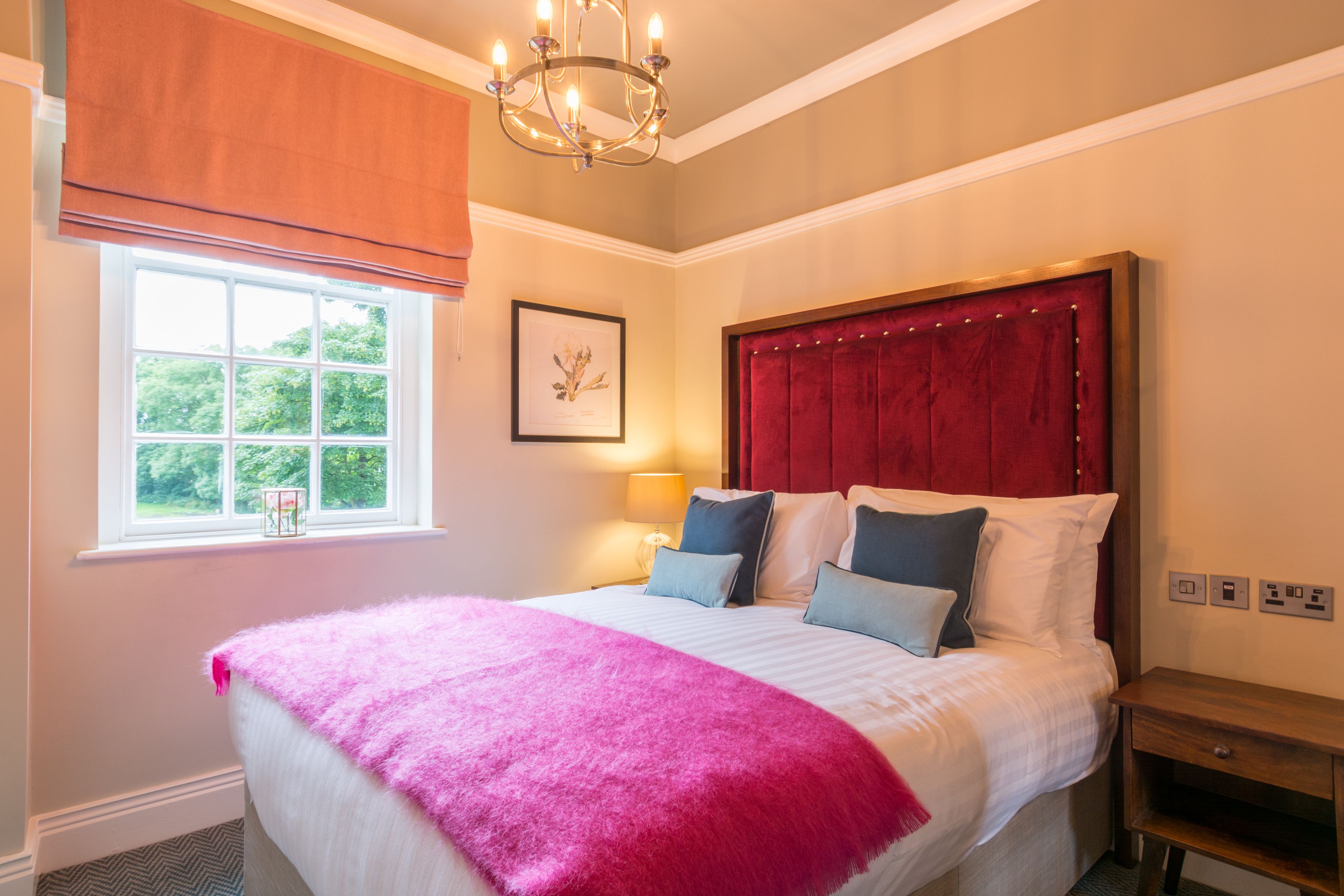 A bedroom with a large bed, a window with a view of greenery, a chandelier, and a bedside table.
