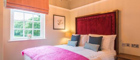 A bedroom with a large bed, a window with a view of greenery, a chandelier, and a bedside table.
