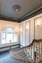 Una gran escalera con barandilla blanca, un candelabro y un amplio ventanal con vistas a la vegetación.