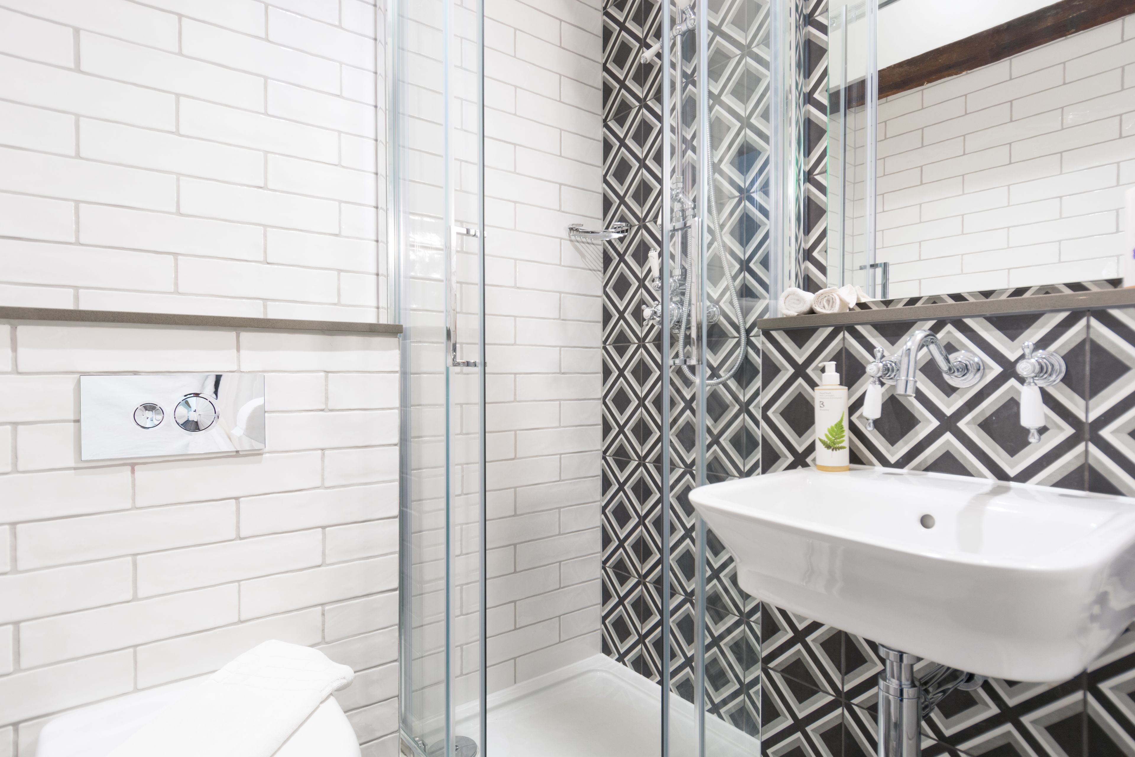Un baño moderno con azulejos blancos tipo metro, mampara de ducha de vidrio, pared con diseño en blanco y negro y lavamanos blanco con dispensador de jabón.