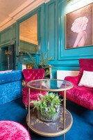 Una habitación color turquesa con un sofá azul, un sillón de terciopelo rojo y una mesa de centro de vidrio con una planta.