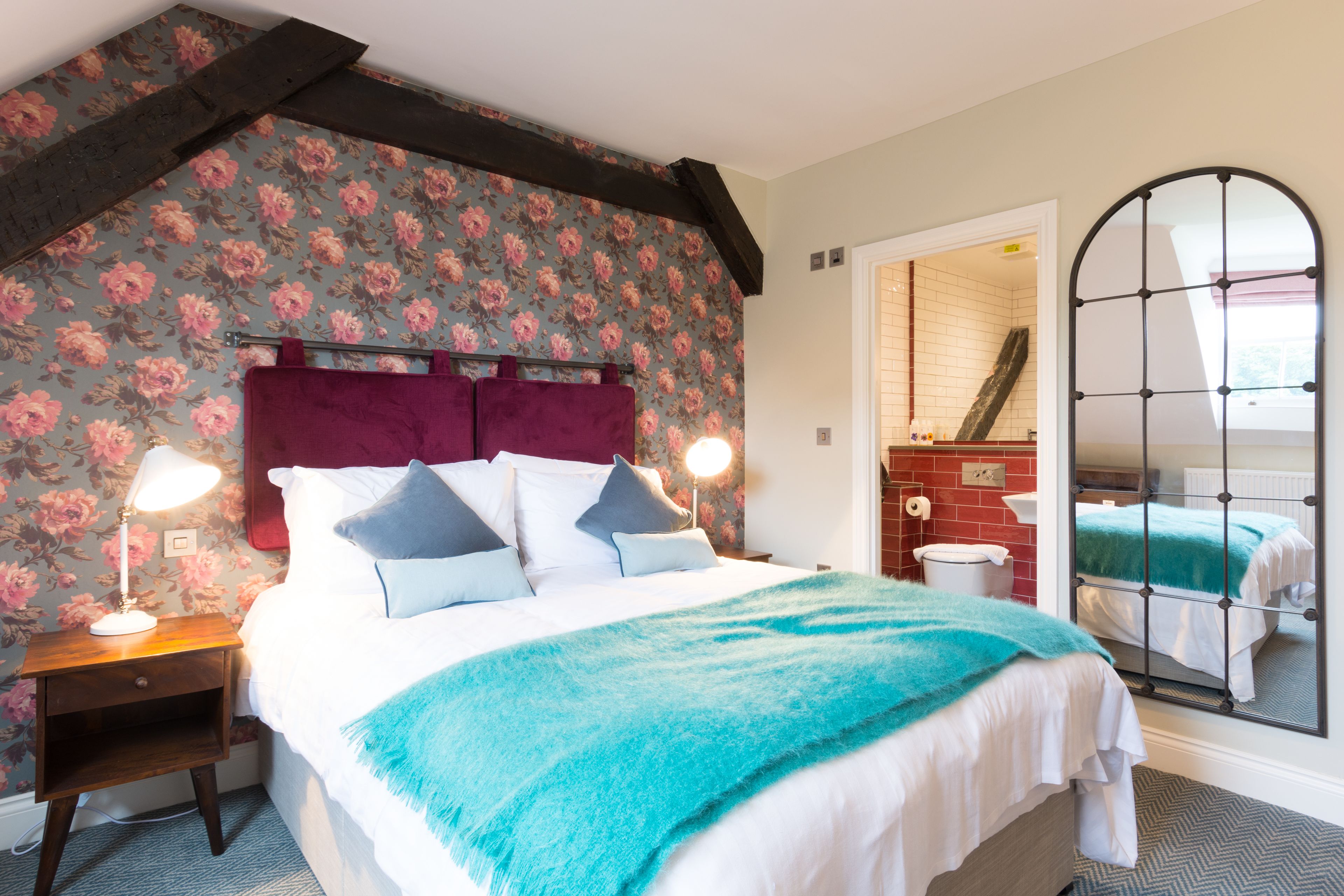 A bedroom with a large bed, floral wallpaper, a wooden headboard, and a mirror with a grid design.