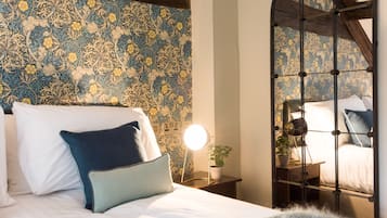 Een slaapkamer met behang met een patroon, een bed met wit en blauw beddengoed, een nachtkastje met een lamp, en een bijzettafeltje met een plant.