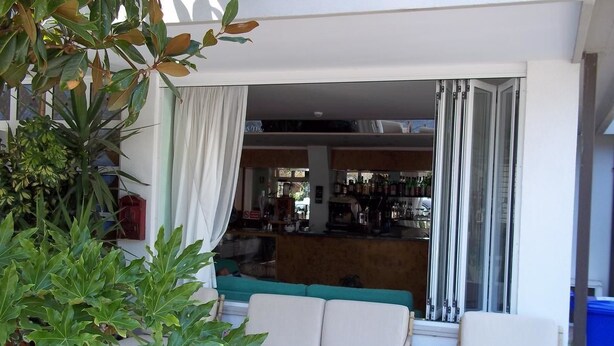 Uma área de pátio com sofá e cadeiras, um bar visível pela janela e uma planta grande em primeiro plano.