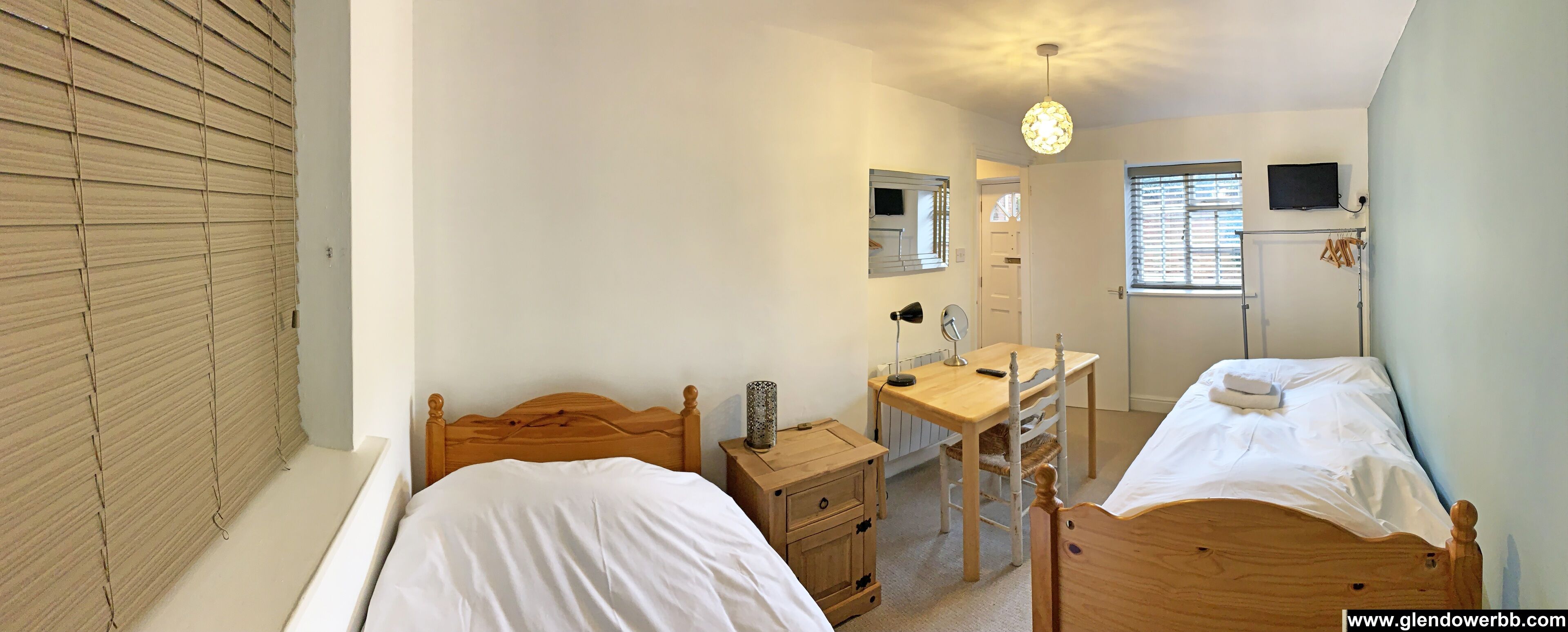 Une chambre à coucher avec deux lits en bois, un bureau en bois avec une chaise, une table de chevet, un miroir, un luminaire au plafond et une fenêtre avec des stores.
