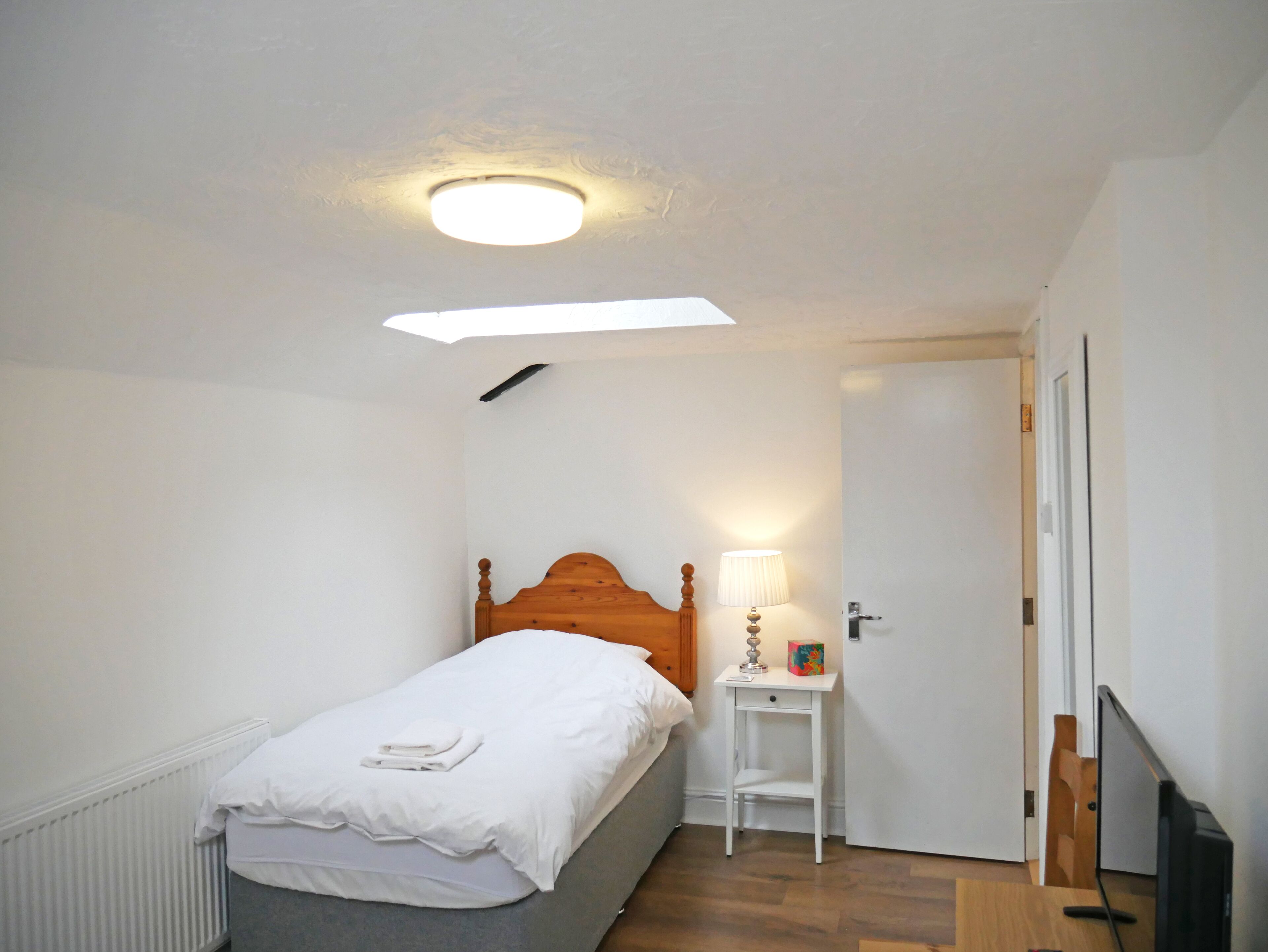 Una habitación pequeña y limpia con una cama individual, una mesita de noche con lámpara y una puerta que da a otra habitación.