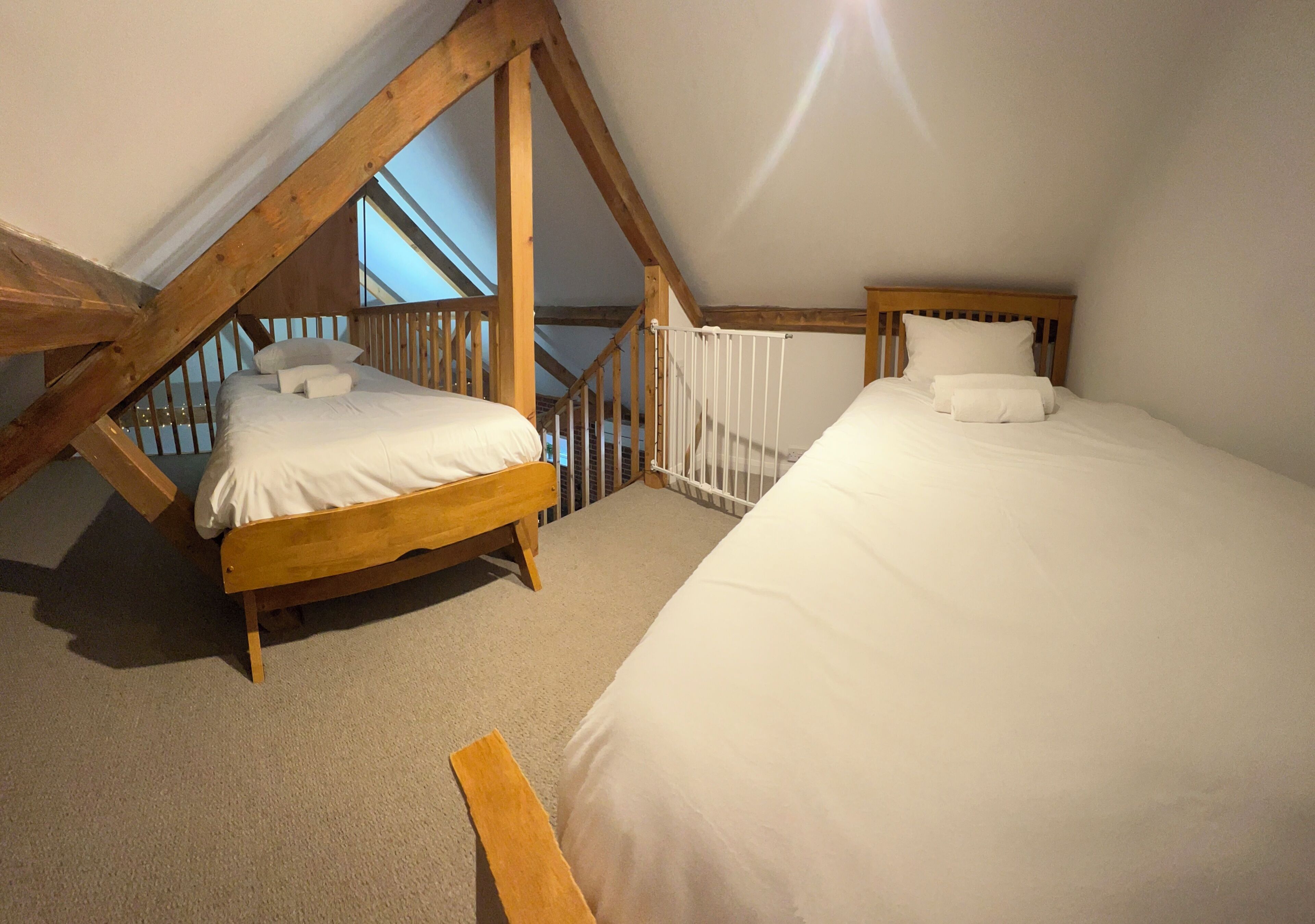 A bedroom with two beds, wooden beams, a white railing, and a view of the sky through a window.