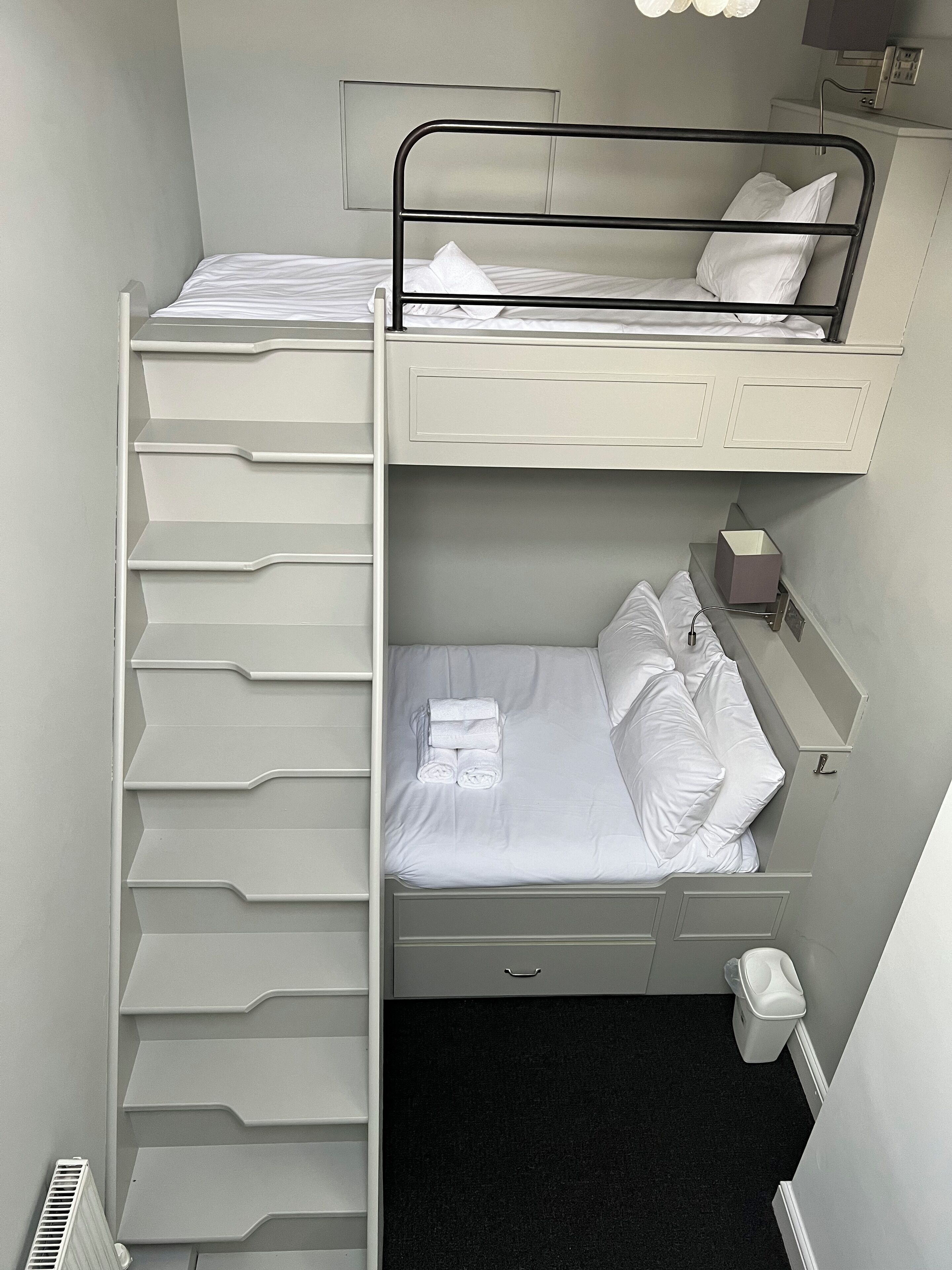 A bunk bed with a pull-out desk and storage drawers, a bedside table with a lamp, and a trash bin.