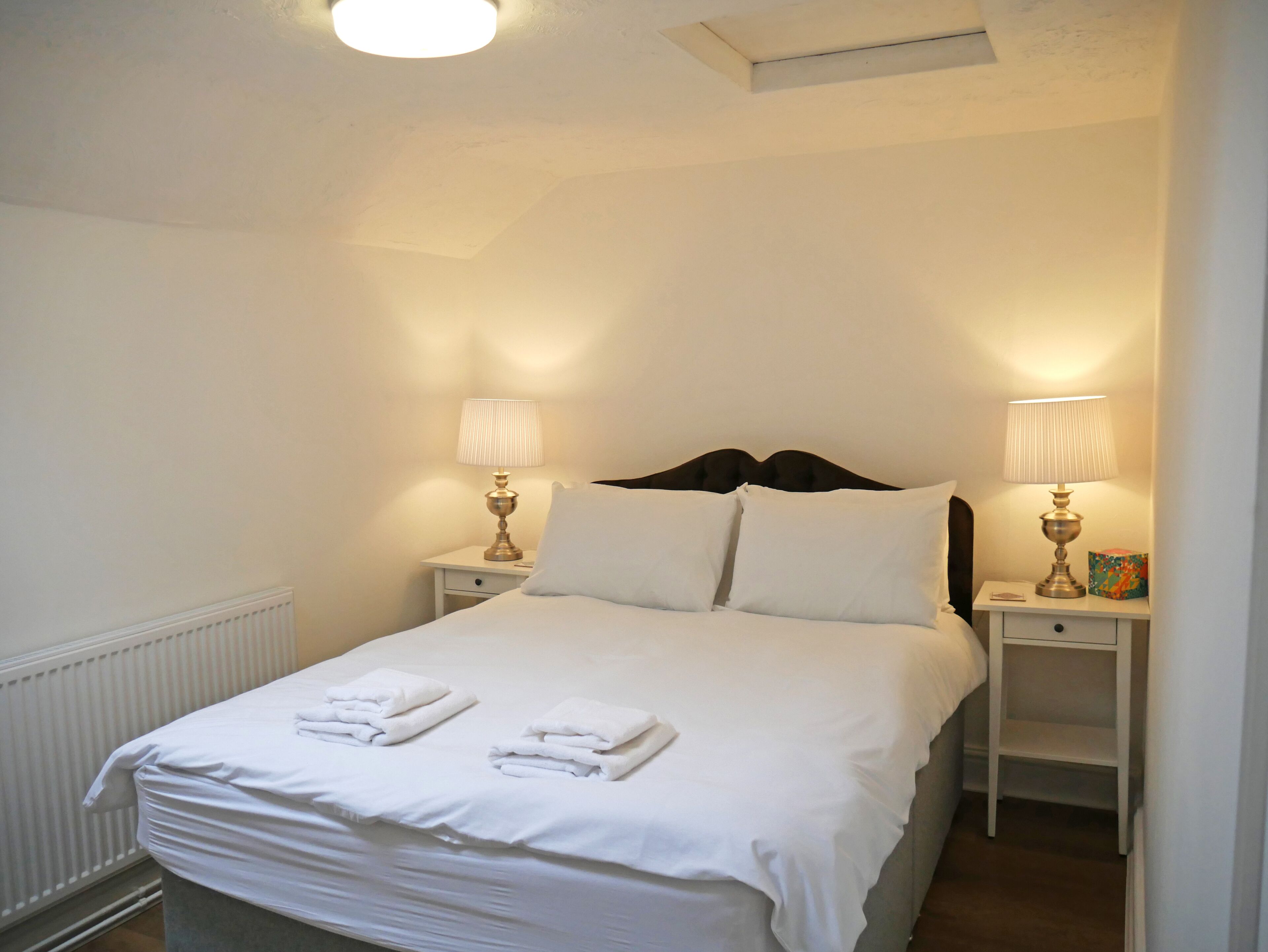 A neatly made bed with white linens and towels, two bedside tables with lamps, and a radiator under the bed.