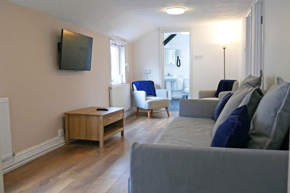 Una sala de estar con un sofá gris, una pequeña mesa de madera, un sillón blanco y un televisor montado en la pared.