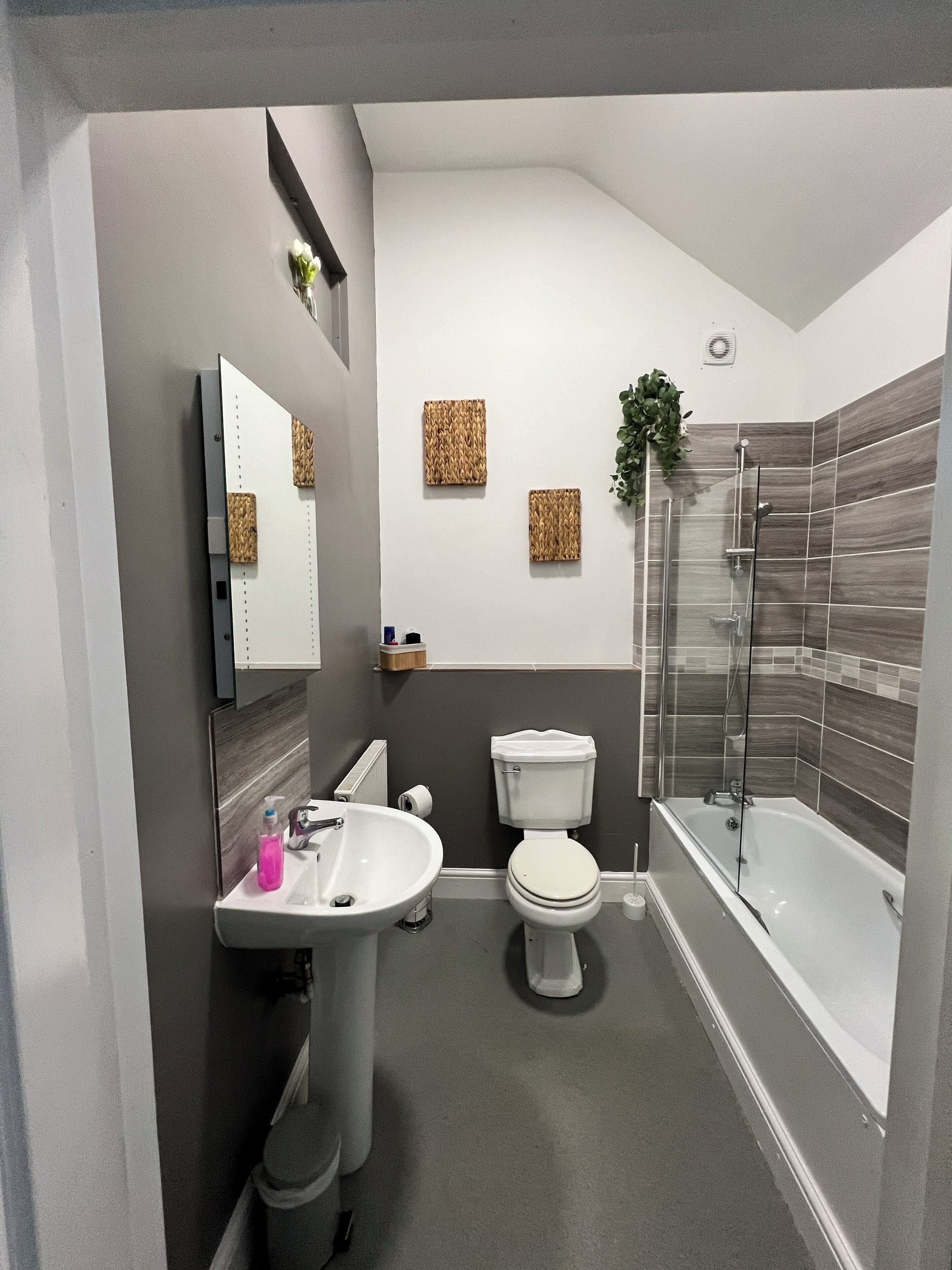 A modern bathroom with a white sink, toilet, and bathtub. There are decorative woven panels on the wall and a small plant.